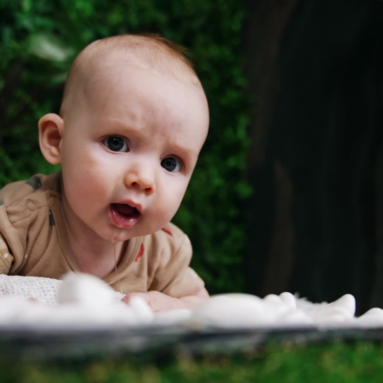 Nydelig barn som ligger på magen og ser seg rundt med overraskelse. Bedårende kaukasisk baby med store øyne. Nærbilde. Grøntområde i bakgrunnen