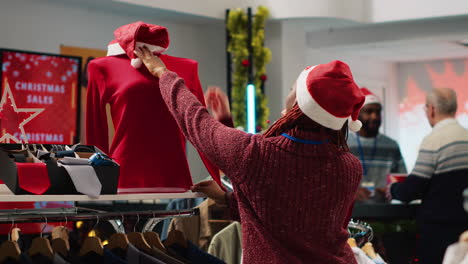 african american retail assistant wearing santa hat helping asian client browsing through festive clothing in christmas shopping store during winter holiday season. employee assisting customer