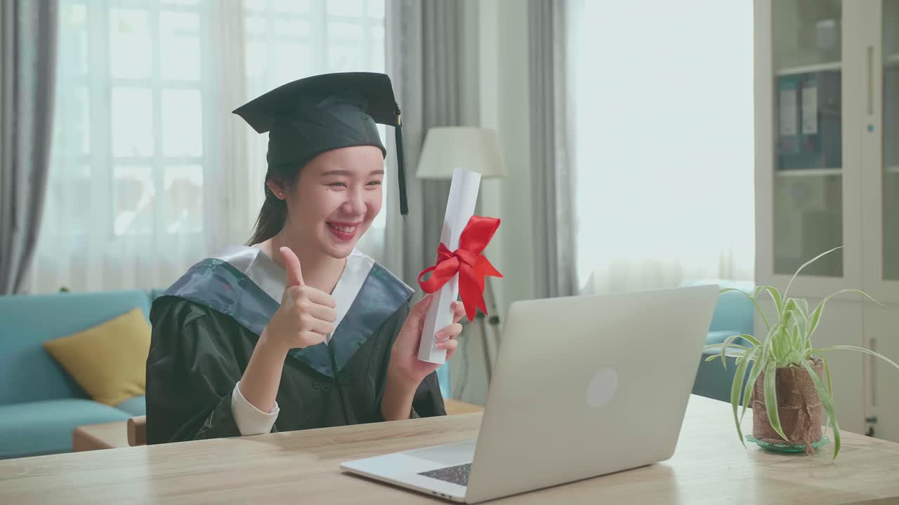 Excited Asian Woman Thumb Up And Showing Off A University Certificate To The Family During An Online Video Call. Pretty Female Graduate Wearing A Graduation Gown And Cap Sitting On The Living Room