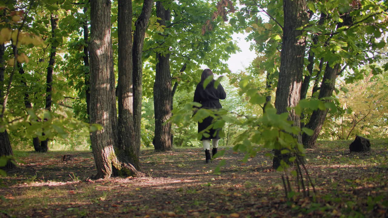 Back view of forest visitor strolling under autumn canopy, sipping warm beverage while looking around, soft sunlight flickering through leaves, coat and boots moving along quiet path, calm mood
