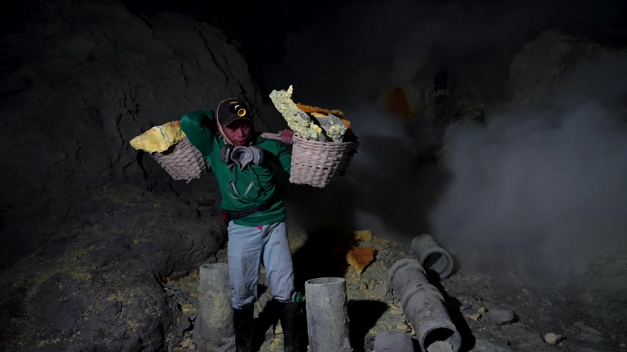 Un minatore di zolfo solleva un cesto pieno sulla spalla tra i fumi vulcanici all'interno del Kawah Ijen in un'inquadratura statica a mano.