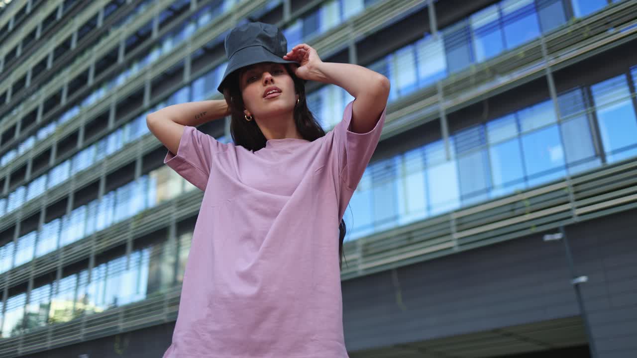 Giovane donna in posa con un cappello da pescatore di fronte a un moderno edificio urbano.