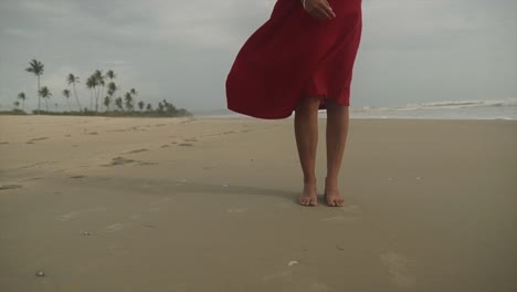toma cinematográfica de una modelo de moda india con un vestido rojo de pie en una playa de arena en goa, india en un día ventoso