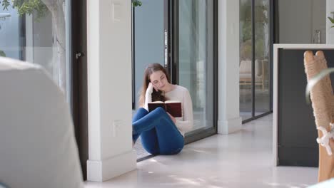 Glückliche-Kaukasische-Frau,-Die-Auf-Dem-Boden-Sitzt-Und-In-Zeitlupe-Ein-Buch-Liest