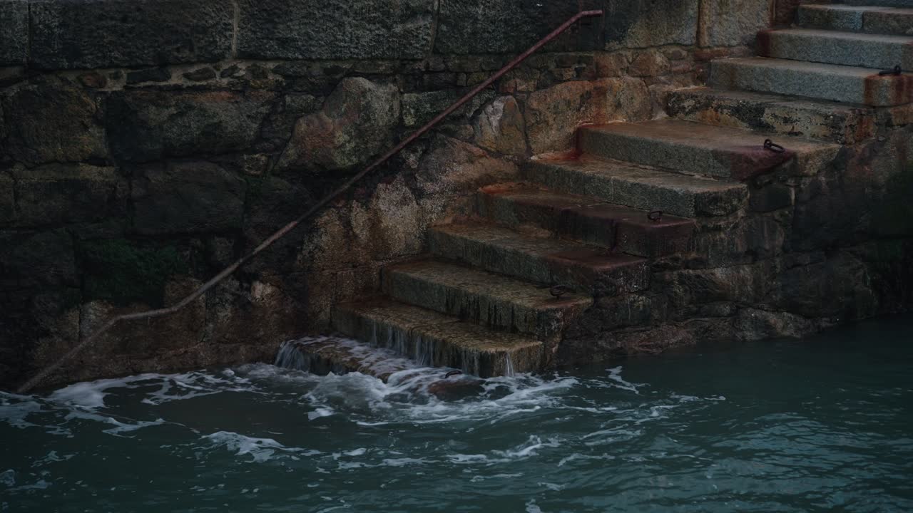 A locked-off shot of ocean waves crashing against weathered stone steps at Colliemore Harbour, Dublin, Ireland. Water splashes against the textured seawall under moody lighting.