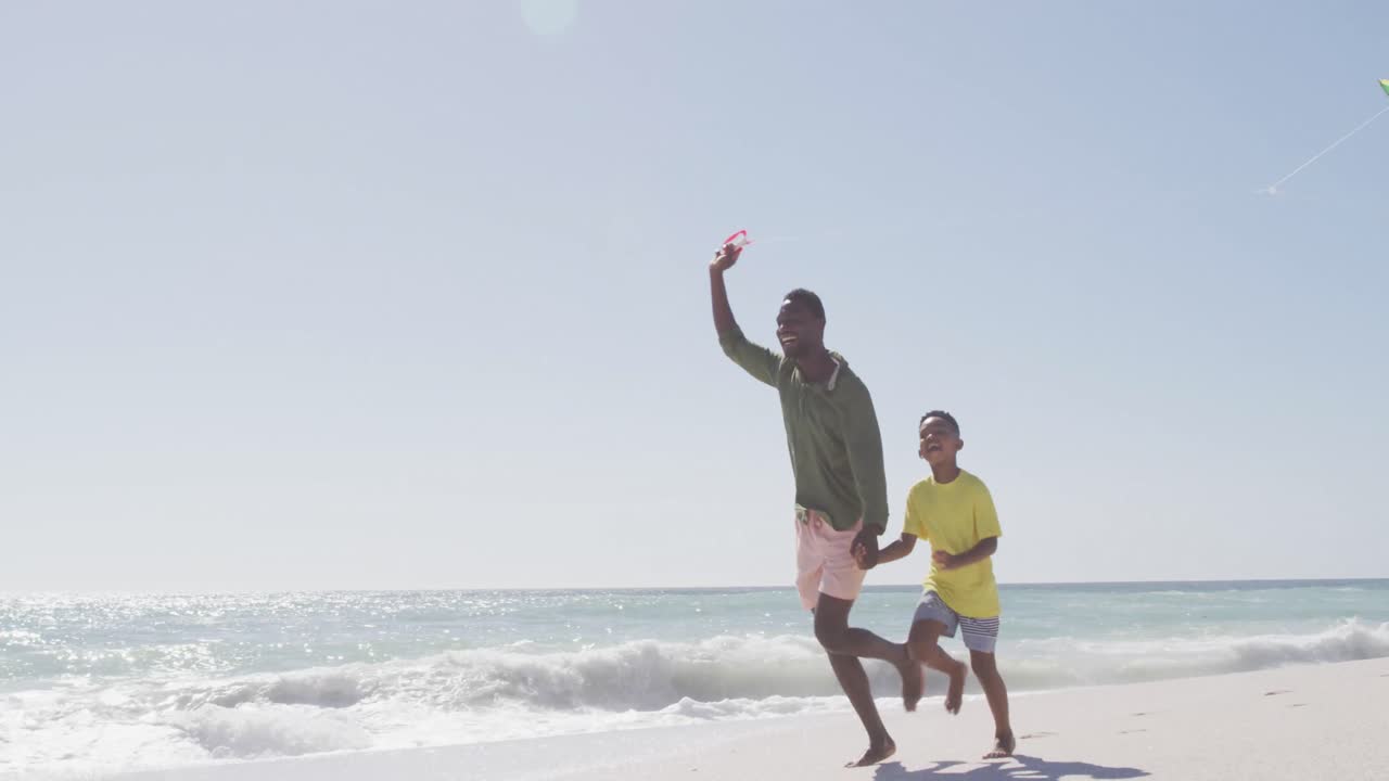 Smiling african american father with son flying kite on sunny beach