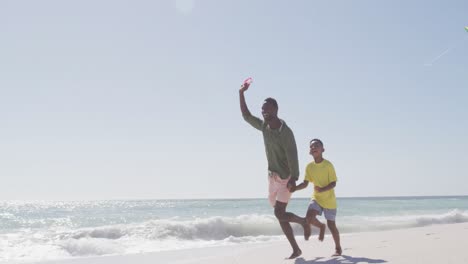 padre afroamericano sonriente con hijo volando cometa en una playa soleada