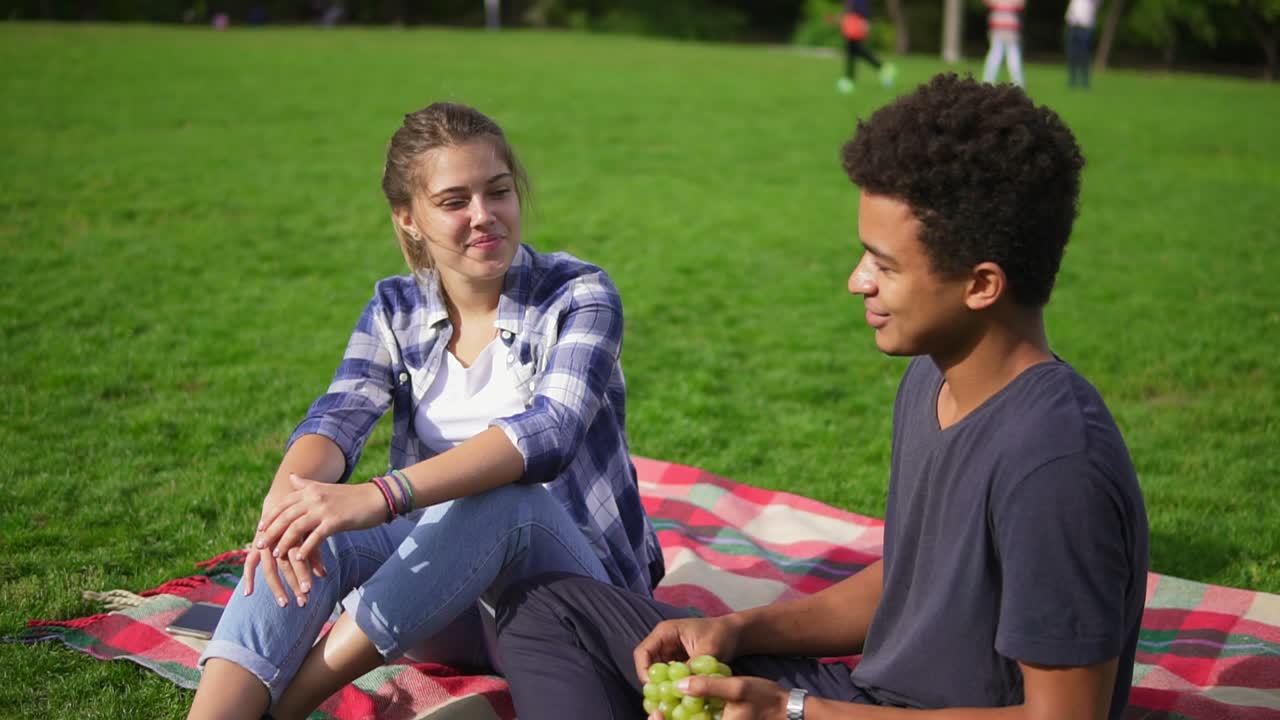 amigos lindos sentados en la hierba y hablando en el parque en un día de verano. chico guapo sosteniendo uvas jugosas en sus manos y diciendo
