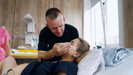 caring man wiping the face of his wife lying in the hospital bed. caucasian couple is preparing for baby delivery.
