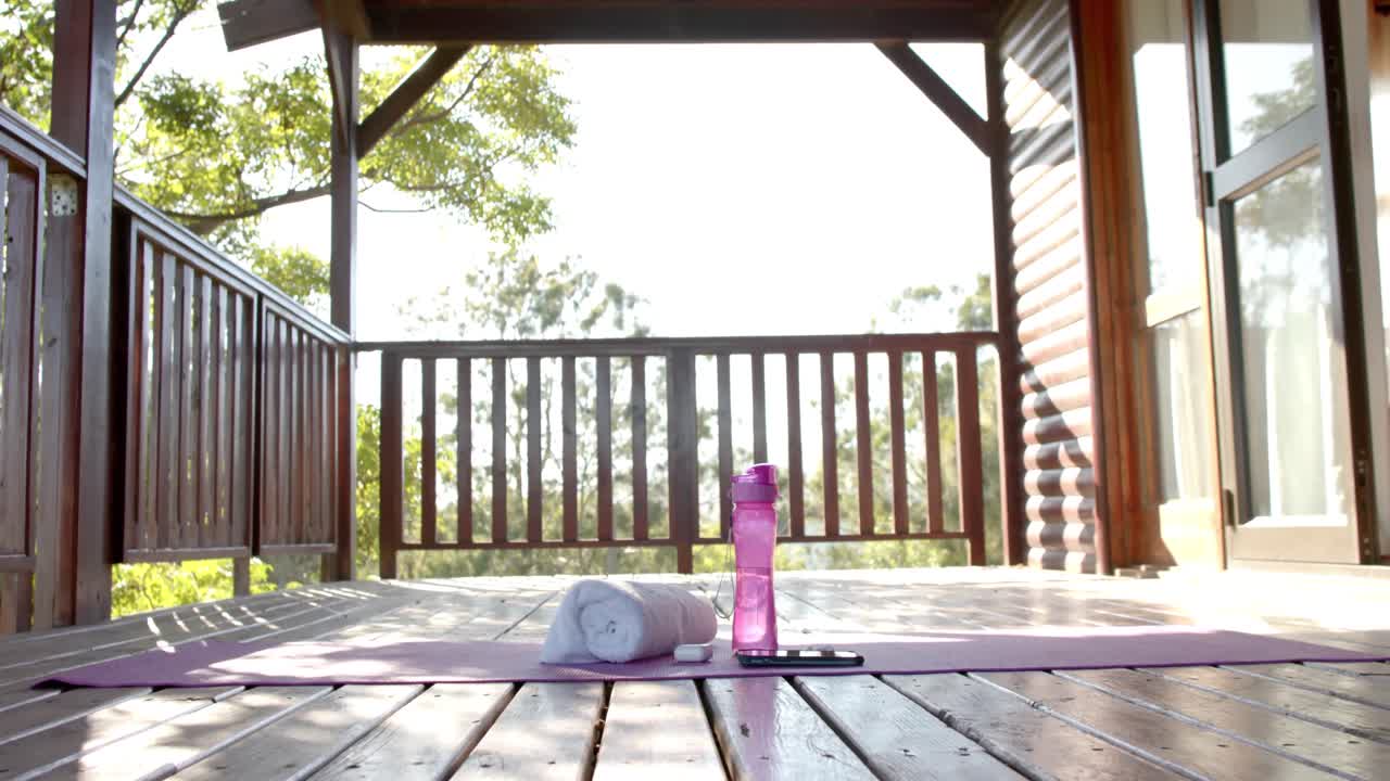 Håndklæde, vandflaske, smartphone og høretelefoner liggende på yogamåtte på solrig terrasse, slow motion