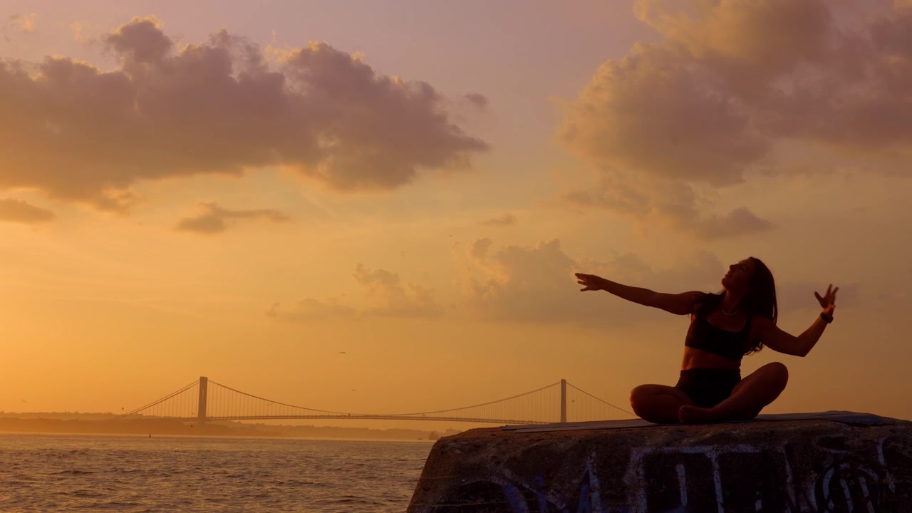 Kvinde sidder i meditationsstilling under den gyldne time nær New Yorks havn. Kvindelig figur i skrædderstilling på en stenblok. Varm aftenbelysning, skyer og en stor hængebro i baggrunden