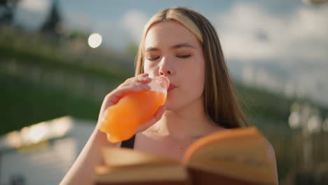 mujer sentada al aire libre, sosteniendo un libro en una mano mientras toma un sorbo de una botella de jugo con la otra, el libro está ligeramente borroso mientras continúa leyendo, el fondo está borroso