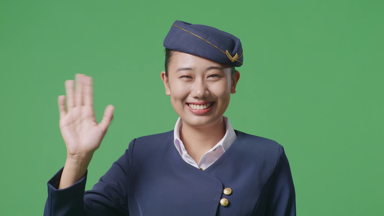 primer plano de una azafata asiática sonriendo, agitando la mano y diciendo adiós mientras está de pie en el estudio de fondo de la pantalla verde
