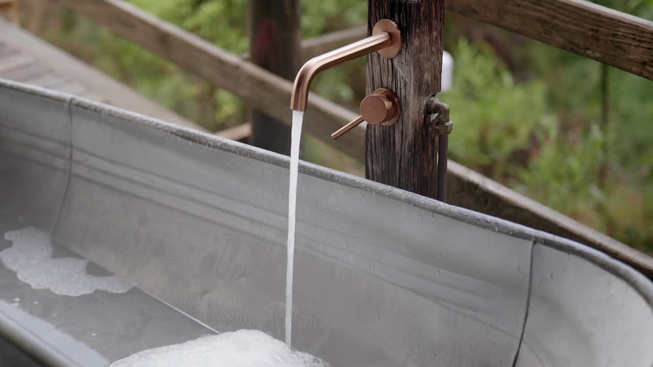 Slow motion gimbal shot of copper tap flowing into outdoor metal bathtub creating bubbly foam with wooden railing and greenery