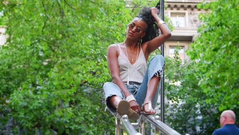 trendy young black woman wearing camisole and side stripe jeans, sitting on handrail in green city street