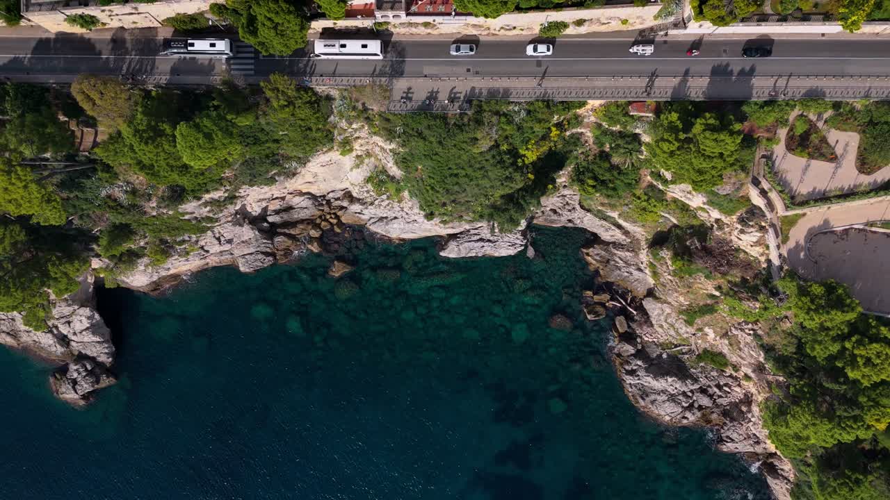 Bird’s-eye aerial drone captures Dubrovnik’s cliffside coast, featuring a winding road along the edge with cars passing by, terracotta-roofed buildings below, and the deep blue Adriatic Sea