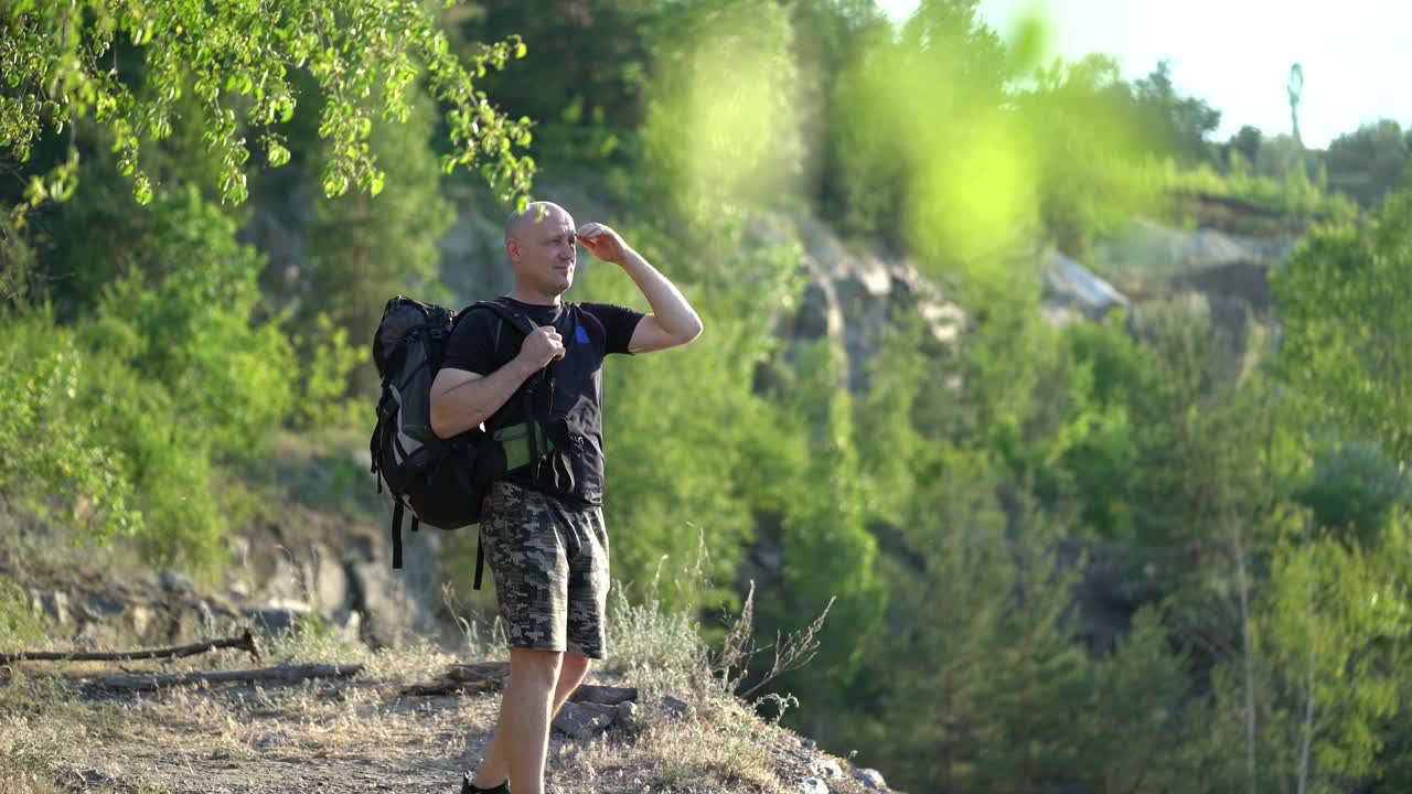 Hiker male looks at the beautiful landscape on the natural green background. A tourist man with a rucksack standing on the hill and admires the nature in a sunny day