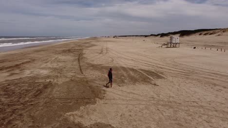 Slow-motion-shot-of-an-old-man-walking-on-the-sand-in-front-of-the-beach-water-waves-on-a-natural-background-at-Mar-de-las-pampas,-Argentina