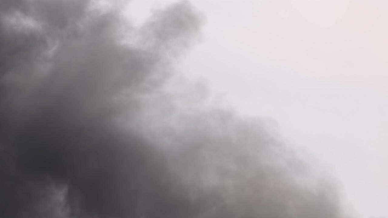 Thick, dark smoke swirls dynamically against a light sky, creating a dramatic and moody atmosphere