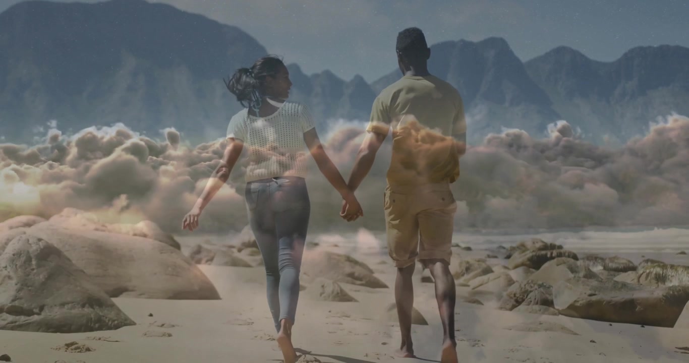 Una coppia in viaggio cammina su una spiaggia rocciosa, mentre il loro passo intensifica una sovrapposizione di nuvole che si dirige verso le montagne.