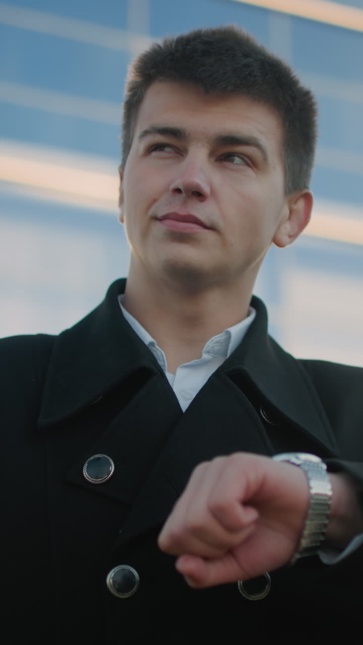 hombre con ropa negra con sonrisa sutil revisa el reloj de muñeca mientras mira a la distancia, exuda confianza y compostura, el fondo presenta un edificio de vidrio moderno que refleja la luz solar del hombre