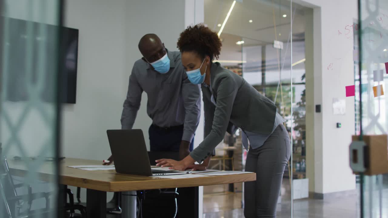 Diverse male and female business colleagues in face masks discussing, using laptop in office