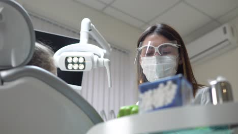 Friendly-female-dentist-turning-on-the-lights-and-taking-her-instruments-before-doing-routine-check-up-of-a-cute-little-girl.-Shot-in-4k