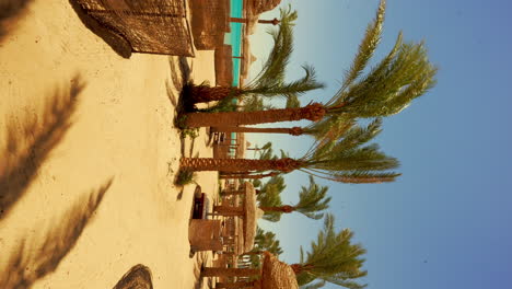 Palmeras-Se-Balancean-En-El-Resort-Frente-A-La-Playa-De-Hurghada,-Video-Vertical-De-Cabañas-Con-Techo-De-Paja