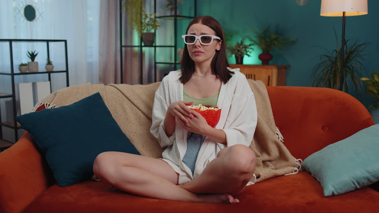 Donna seduta sul divano che mangia popcorn e guarda un'interessante serie TV o una partita sportiva online da casa.