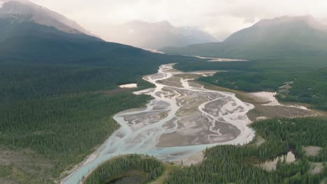 Aerial-truck-left-of-river-trail-surrounded-by-dense-pine-tree-forest-and-Canadian-Rockies-at-Banff-National-Park,-Alberta,-Canada