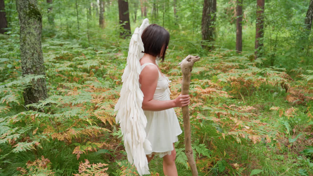 Young woman wearing white dress and large feathered wings walks through lush green forest with staff, gently touching ferns and flowers, embodying ethereal goddess energy in serene natural setting