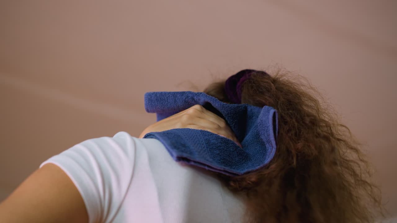 Side view of girl with tied hair cleaning sweat from neck using blue towel after intense workout showing recovery effort and relief inside fitness studio environment with determination expression