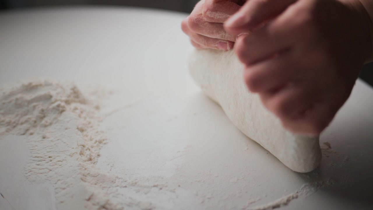 Mani impastano impasto bianco su una superficie infarinata. Il primo piano cattura il movimento tattile e la consistenza. Focus sul processo dettagliato di preparazione del pane o della pasticceria. Ideale per progetti culinari, alimentari o a tema artigianale.