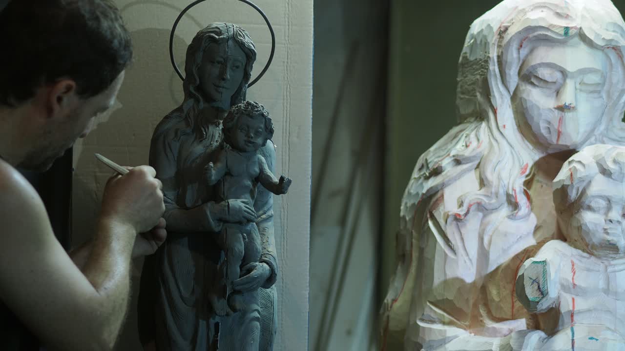 An artisan sculpts a clay model of the Virgin and Child, using it as a reference for a larger wooden statue in progress, showing the process of religious art creation