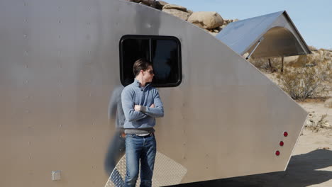 a handsome young white man traveler leaning against a metal teardrop travel trailer tiny house on his solo road trip