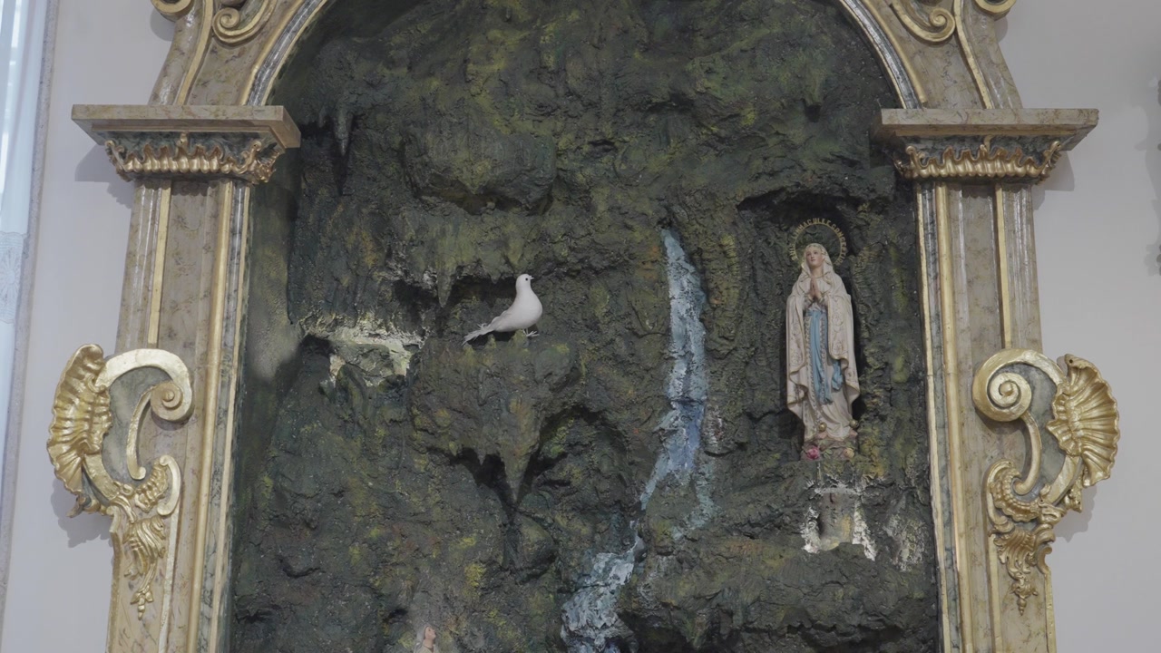 Detail of a sacred indoor grotto featuring a statue of Our Lady of Lourdes, a white dove, and rock textures.