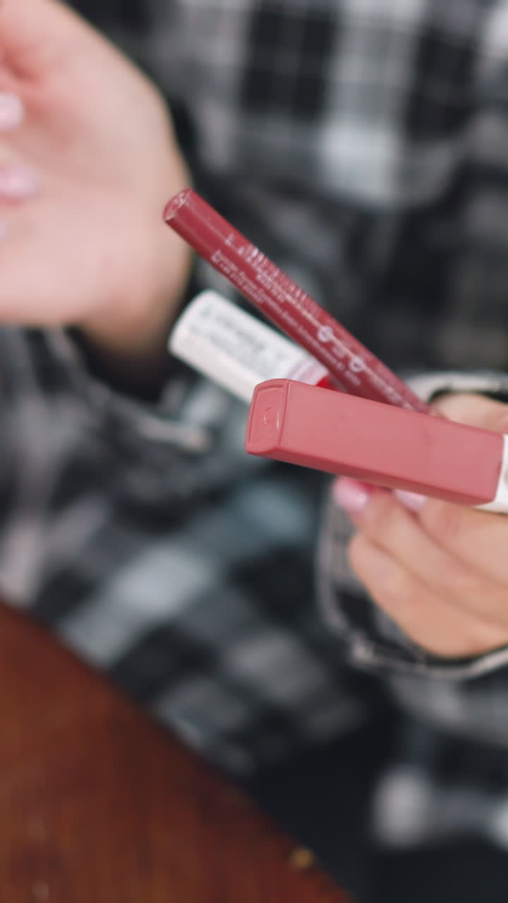Primer plano de unas manos revisando la etiqueta del envase de un lápiz de labios; una mujer con camisa de cuadros examina el perfilador de labios y la caja, se aprecia una manicura cuidada, inspección minuciosa del tono y los ingredientes, luz acogedora sobre la mesa.