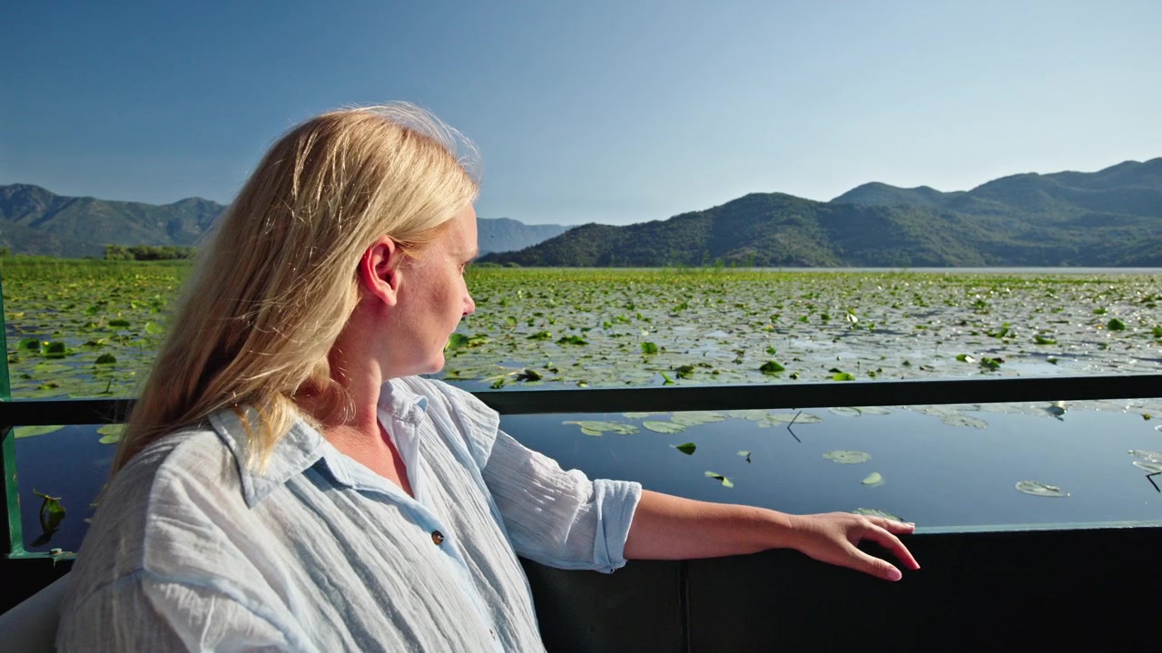 Middle aged woman enjoying a peaceful boat trip across Skadar Lake National Park in Montenegro. She admires water lilies and mountain landscapes