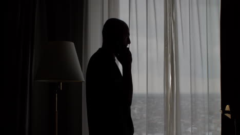 Silhouette-of-bald-man-walking-towards-the-window-with-the-curtains-closed,-talking-on-a-mobile-phone,-the-concept-of-privacy-and-anonymity