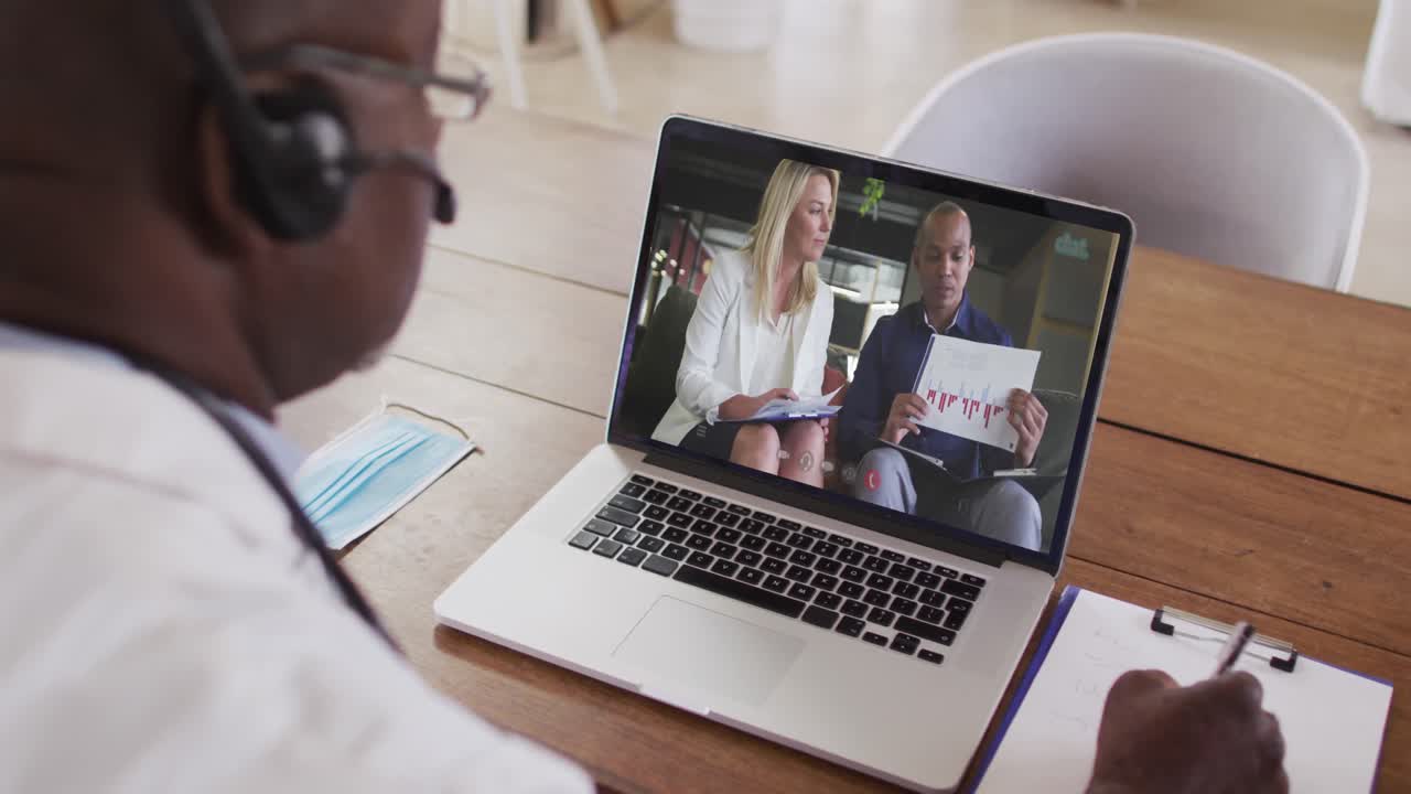 Afroamerikansk mandlig læge med headset på, der tager noter under et videoopkald på sin bærbare computer
