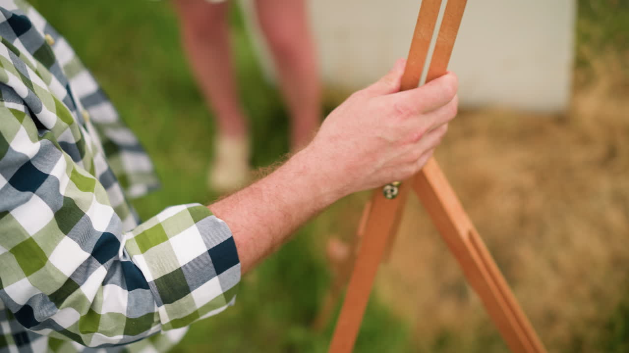 A close-up of a person's hand, wearing a plaid shirt, adjusting a wooden tripod in a grassy field. The surrounding area is blurred, with a woman's leg visible in the background