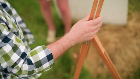 un primer plano de la mano de una persona, con una camisa a cuadros, ajustando un trípode de madera en un campo cubierto de hierba. el área circundante está borrosa, con la pierna de una mujer visible en el fondo