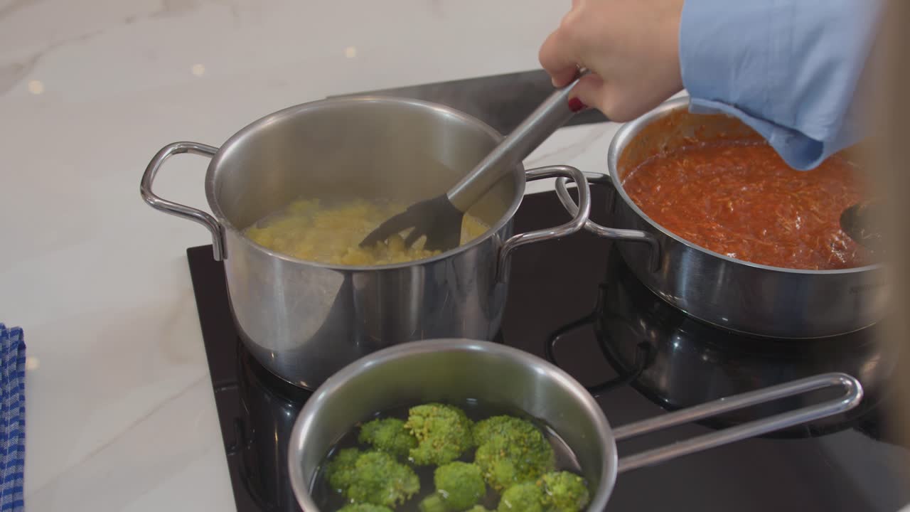 Uma mulher está cozinhando brócolis em uma panela. A panela está em um fogão. A mulher está mexendo o brócolis com uma espátula.