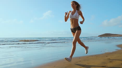 Beautiful-sportive-woman-running-along-beautiful-sandy-beach,-healthy-lifestyle,-enjoying-active-summer-vacation-near-the-sea.-SLOW-MOTION-STEADICAM.