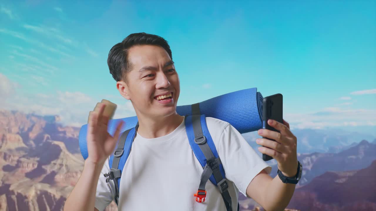 Close Up Of Asian Male Hiker With Mountaineering Backpack Waving His Hand And Pointing Back While Having A Video Call On Smartphone While Traveling At The Top Of Mountain