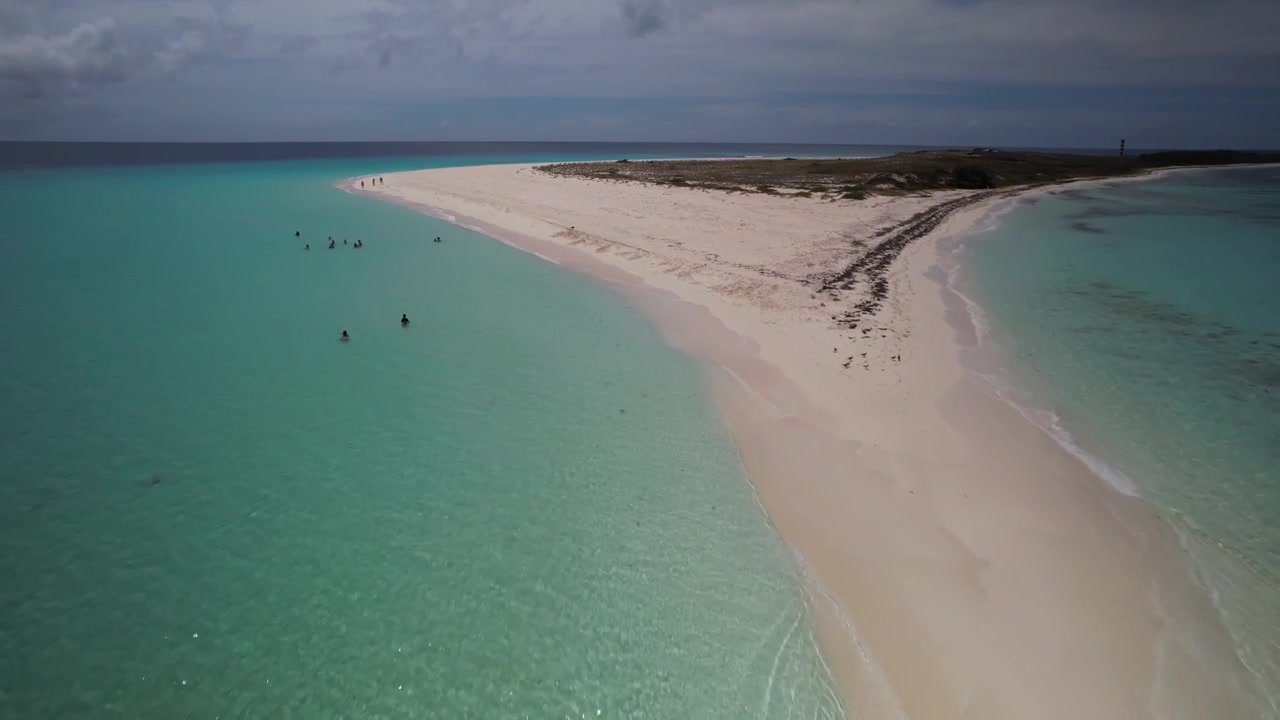 High-angle drone view gliding over crystal-clear turquoise waters along a remote sandbar where people swim and walk in paradise.
