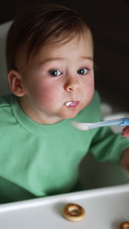 sweet toddler eating yoghurt from spoon given by his mom. grey-eyed child sits at feeding table and looking up at camera with interest. high angle view. vertical video