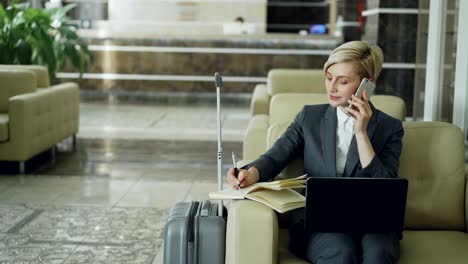 pan shot of blonde busy businesswoman sitting in armchair in hotel lobby talking mobile phone, writing in notepad and using laptop computer. business, travel and people concept