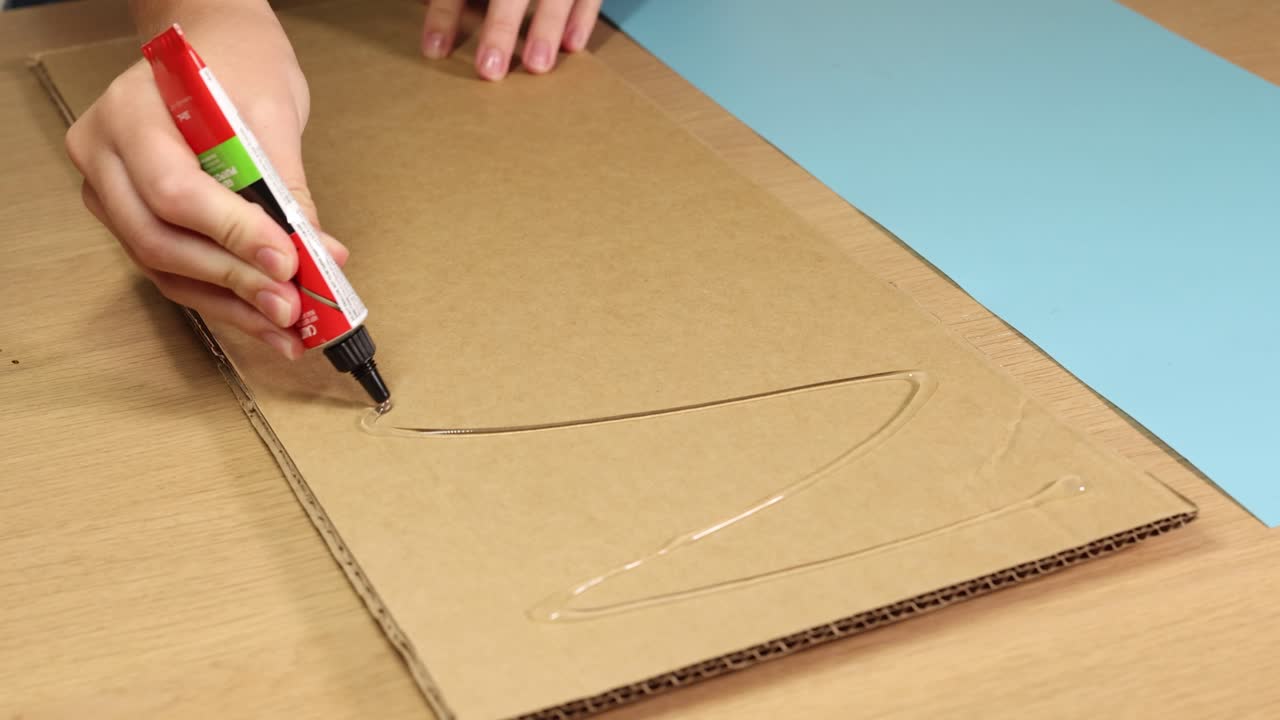 A hand applies glue to a cardboard sheet on a wooden table under bright lighting, showcasing a crafting process