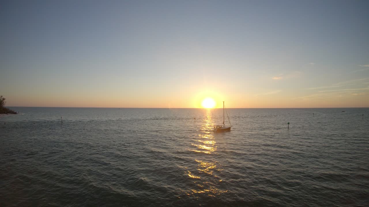 A boat crossing the sea in front of the sun setting over the horizon. Drone footage moves backwards captur8ng this beautiful scene and shows some of the beach and surrounding trees.